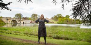 Hannah with outstretched arms in front of a lake with a bridge.
