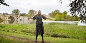 Hannah with outstretched arms in front of a lake with a bridge.